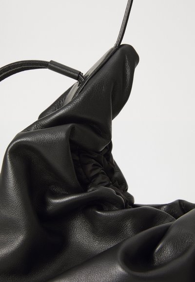 Minkštas, elastingas juodas odinis rankinukas su susiraukšlėjimais ir plona petneša, prieš neutralų šviesų foną.