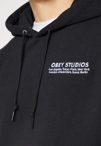 Černá mikina s kapucí z bavlněné směsi, s přední kapsou, nastavitelnou šňůrkou na kapuci a vyšívaným logem s názvy měst v bílé barvě.