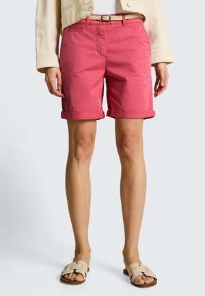 Mujer con pantalones cortos color coral rosa con dobladillo enrollado y cinturón tejido, chaqueta color crema, camiseta blanca y sandalias beige, de pie sobre un fondo claro.