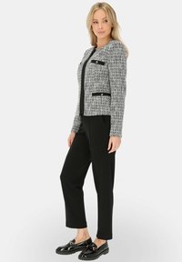 Szara i czarna marynarka z tkaniny tweedowej o krótkim kroju, z dwoma czarnymi dodatkami przy kieszeniach, zestawiona z czarnymi spodniami i błyszczącymi czarnymi butami.