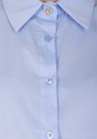 Camicia azzurra chiara con colletto a punta, bottone circolare decorativo al collo e bottoni standard lungo la parte anteriore. Texture liscia.