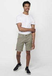 Jonge man die staat en glimlacht, gekleed in een wit t-shirt, olijfgroene shorts, zwarte sokken en zwarte sneakers met witte accenten.