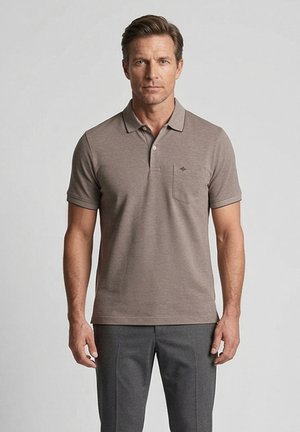 Mann trägt ein kurzärmeliges Poloshirt in Taupe mit Brusttasche und graue Hose, steht vor einem schlichten grauen Hintergrund.