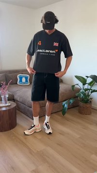 Osoba u crnoj majici McLaren Formula 1 tima, crnoj kapi, kratkim hlačama i tenisicama stoji u dnevnoj sobi s kaučem, biljkom i drvenim podom.