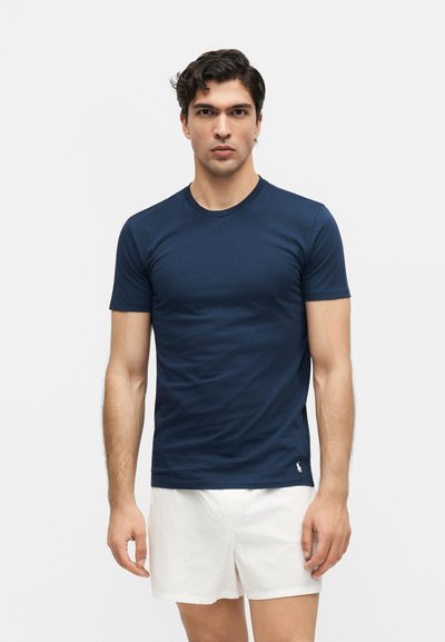 Νέος με σκούρα μαλλιά φοράει εφαρμοστό κοντομάνικο μπλουζάκι σε μπλε ναυτικό χρώμα και λευκά σορτς, όρθιος μπροστά σε απλό φωτεινό φόντο.