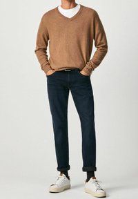 Homme portant un pull marron à col en V, un jean bleu foncé retroussé aux chevilles, des chaussettes noires et des baskets blanches, debout les mains dans les poches.