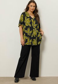 Chemise noire avec motif de feuilles vertes, manches courtes, décolleté en V, coupe ample, associée à un pantalon large noir et à des bottes à talons noires.
