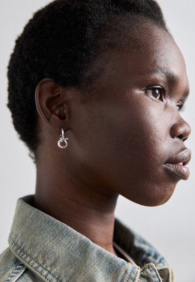 Silver hoop earrings featuring a small lever closure and a circular accent. The model has short, dark hair and wears a denim jacket.