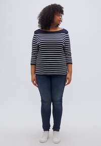 Μπλούζα με μακριά μανίκια σε ναυτικό μπλε και λευκές ριγέ, με φαρδύ ντεκολτέ, συνδυασμένη με σκούρα στενά τζιν και λευκά αθλητικά παπούτσια. Σγουρά μαλλιά, χέρια στα πλευρά.