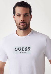 White cotton t-shirt featuring a black and mint green "GUESS EST. 1981" graphic print on the front. Short sleeves and crew neck design.