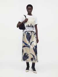 Femme portant un t-shirt graphique blanc, une jupe fleurie beige et bleue brillante, des chaussettes noires avec des talons blancs, et tenant un sac à épaule noir.