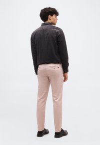 Άνδρας που φοράει μαύρο πουκάμισο με μακριά μανίκια, ανοιχτό ροζ παντελόνι και μαύρα παπούτσια, στέκεται με την πλάτη σε ένα απλό ανοιχτό φόντο.