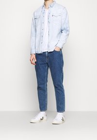 Camicia di jeans azzurro chiaro indossata sopra una maglietta bianca, abbinata a jeans blu medio e scarpe da ginnastica bianche con un logo scuro.