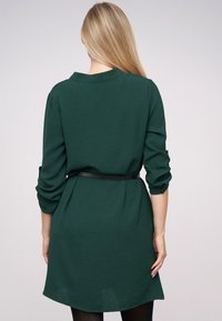 Grünes Hemdkleid aus weichem Stoff, mit Kragen, langen Ärmeln und einem schwarzen Gürtel in der Taille. Glatte Textur, abgerundeter Saum.