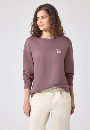 Femme aux cheveux bouclés porte un sweat mauve avec un petit motif de cerise rose, assorti à un pantalon crème taille haute, debout calmement.