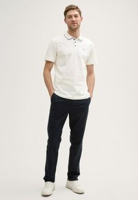 Polo bianco con collare blu scuro e dettagli in bottone, abbinato a pantaloni blu scuro e sneakers bianche. Tessuto morbido e vestibilità rilassata.