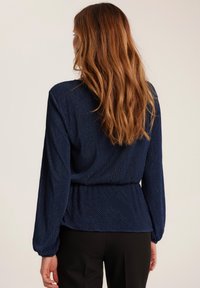 Granatowa bluzka z pionowym żebrowanym wzorem, długimi rękawami i marszczeniem w talii, w zestawieniu z czarnymi spodniami. Widok z tyłu.