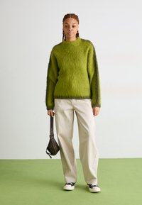 Πράσινο φλας πουλόβερ με στρογγυλή λαιμόκοψη και αντίθετα σκούρα μανίκια, σε συνδυασμό με ανοιχτό καφέ παντελόνι με φαρδιά πόδια και μαύρα αθλητικά παπούτσια.