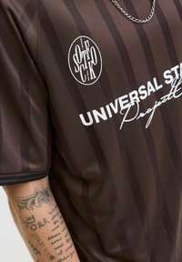 Brown striped jersey with the text "UNIVERSAL STUDIO PROJECT" in white; features a round logo and a contrasting black collar.