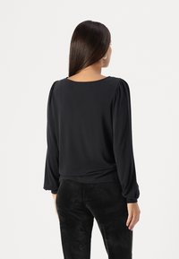 Schwarze Bluse mit langen Ärmeln und dezenten Puffärmeln, runder Halsauschnitt und glattem Stoff. Kombiniert mit engen Hosen in Wildlederoptik.