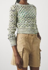 Kobieta ubrana w krótką sweterek o otwartym splotzie w kolorze żółtym i białym, nałożony na zieloną bluzkę, zestawiony z beżowymi szortami cargo z bocznymi kieszeniami.