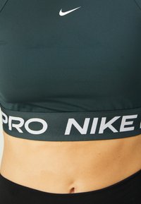 Sportowy biustonosz Nike w ciemnozielonym kolorze, wykonany z gładkiego materiału. Posiada biały logo oraz elastyczny pasek z napisem "PRO" w kontrastowym kolorze. Zbliżenie na pas.