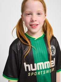 Lächelndes rothaariges Mädchen mit Sommersprossen trägt ein schwarzes und grünes Hummel-Sporttrikot mit Zöpfen vor einem einfarbigen hellen Hintergrund.