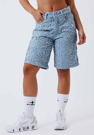 Persona che indossa pantaloncini di jeans azzurri chiari con motivi a spirale in rilievo, calzini bianchi e sneakers bianche Nike, in piedi con una mano in tasca.