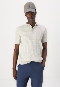 Ljusgrå kabelstickad polotröja med korta ärmar och krage, i kombination med mörkblå byxor. En beige keps fulländar looken.