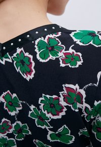 Blusa floral con un fondo oscuro, grandes flores verdes y acentos rojos. Cuello negro adornado con perlas decorativas blancas.