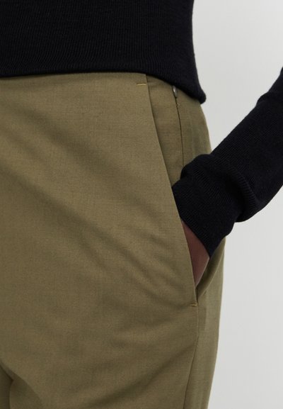 Beige bikses ar gludu tekstūru un sānu kabatu. Jostasvietā ir melns augšējais krāsa un smalka dzeltena šuvju detaļa.