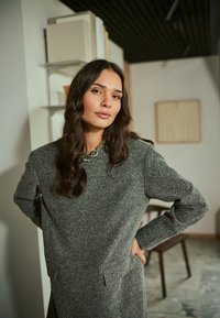 Robe grise texturée avec des manches longues, des poches latérales et un col rond arborant un détail en chaîne décoratif. Design simple et moderne.