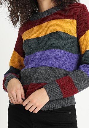 Femme portant un pull tricoté à rayures horizontales dans les couleurs bordeaux, jaune, vert foncé, violet et gris, associé à un pantalon noir.