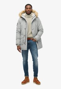 Chaqueta acolchada gris con capucha forrada de pelo, combinada con un suéter de punto ligero, jeans azules y botas de cuero marrones con cordones.