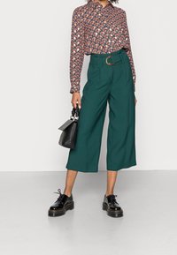 Chemise à boutons imprimée avec un motif de coquillages, pantalon large vert, chaussures noires à semelles épaisses et un petit sac à main noir au design structuré.