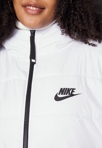Biała kurtka puchowa z wysokim kołnierzem, wyposażona w czarny zamek błyskawiczny i wyraźne czarne logo Nike na przodzie. Gładka, błyszcząca tekstura.