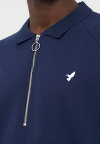 Μπλε ναυτικού με γιακά πουκάμισο με ασημένιο δαχτυλίδι φερμουάρ, μερικώς κλειστό, με μικρό λευκό λογότυπο πουλιού στο στήθος.