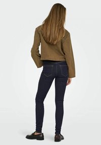 Giacca cropped marrone sopra jeans skinny scuri, con dettagli di cucitura sottili. Le scarpe del modello sono mocassini in pelle scura.
