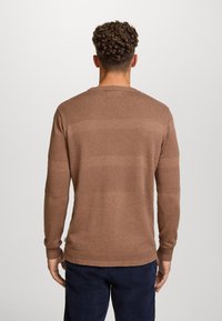Maglione marrone lavorato a maglia con righe testurizzate e scollo rotondo, maniche lunghe e polsini a coste, indossato con pantaloni scuri.