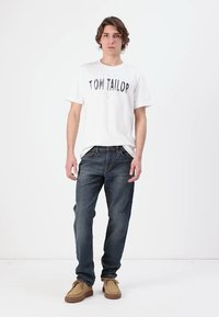 Valge puuvillane T-särk, millel on "TOM TAILOR" trükk, koos tumeda sinise teksapükstega ja beeži seemisnahast kingadega. Mudel poseerib valge tausta ees.