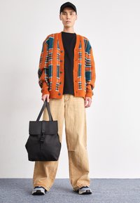 Cardigan lavorato a maglia arancione con motivo teal e bianco, indossato sopra una maglietta nera. Pantaloni beige a gamba larga e uno zaino nero con patta e fibbia.