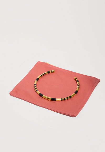 Bracelet ouvert rayé or et noir reposant sur une pochette en tissu corail rose avec des coutures visibles.