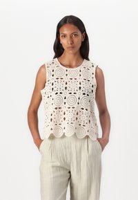 Heklet topp i kremfarge med blomstermønster, ermeløs design, rund hals og bølget kant. Kombinert med lette, plisserte, beige bukser.
