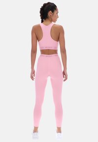 Femme aux cheveux tressés portant un soutien-gorge de sport rose et un legging, avec des baskets blanches, debout de dos sur un fond clair uni.