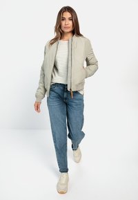 Lys grå bomberjakke med lynlås foran, ribbede manchetter, båret over en stribet top og blå jeans, parret med beige sneakers.
