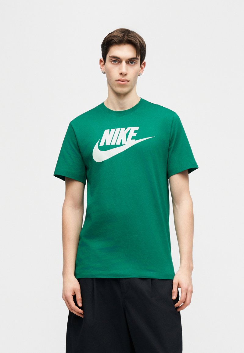 Junger Mann trägt grünes Nike-T-Shirt mit weißem Logo, steht vor einem einfarbigen hellgrauen Hintergrund und blickt nach vorne.