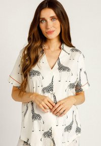 Blouse à manches courtes en tissu crème présentant un motif de girafe noir répété. Détails notables comprenant un décolleté en V et des poignets de manches contrastants.