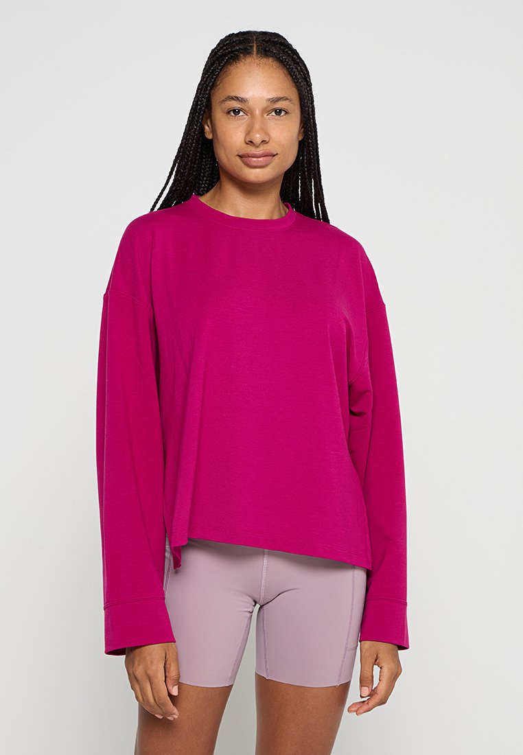 Sudadera oversized de un brillante magenta con mangas largas, cuello redondo y textura suave, combinada con pantalones cortos de ciclismo de un suave color púrpura.