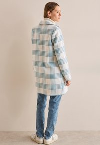 Lång rutiga kappa i vitt med blå och ljus orangea mönster, tillverkad av mjukt material. Matchad med ljusblå jeans och vita sneakers.