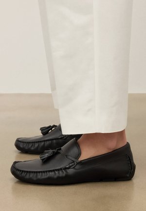 Fekete bőrből készült bojtos mokaszinok, fehér, rövidített nadrággal, sima, semleges színű padlón.